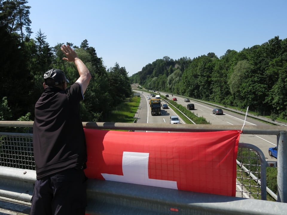 Zu sehen ist ein Mann der winkt, vor ihm hängt eine Schweizer Fahne.