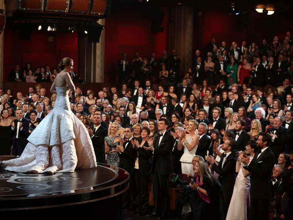 Bühne mit einer Frau in einem weissen Abendkleid mit dem applaudierenden Publikum im Hintergrund.