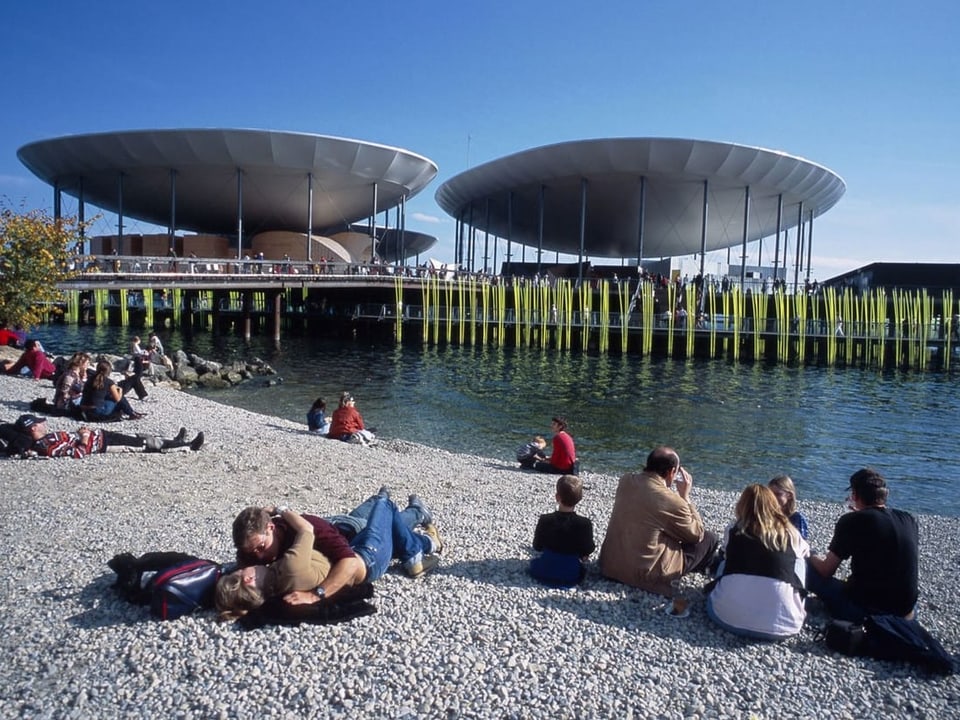 Leute liegen und sitzen am Strand am See, im Hintergrund die Installation der Landessausstellung im See.