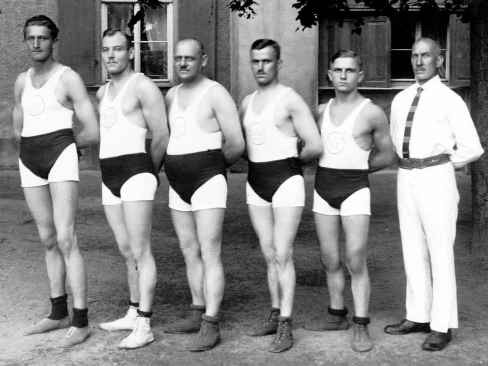 Historical photograph of six male athletes in sports uniforms next to a coach in a white suit