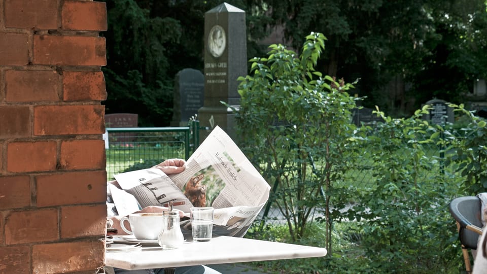Zeitung und Kaffeetasse im Vordergrund, Grabstein im Hintergrund.