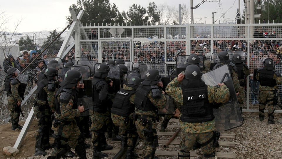 Die mazedonische Polizei, geschützt durch Helme, Westen und Schutzschilder steht zahlreichen Flüchtlingen am Grenzzaun gegenüber.