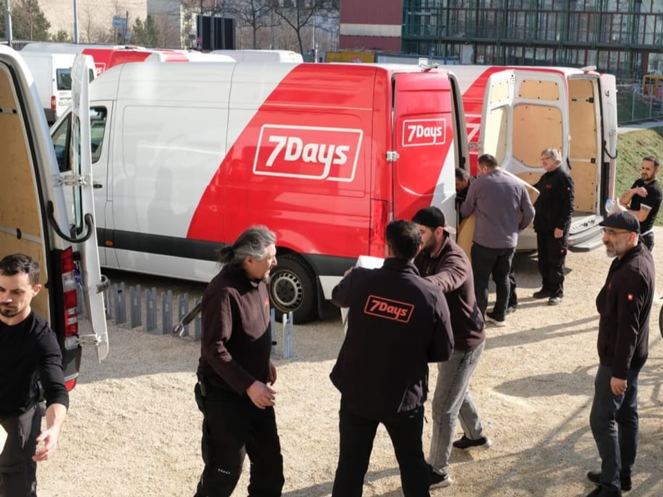 Männer packen Kisten in mehrere Lieferwagen, die nebeneinander geparkt sind.