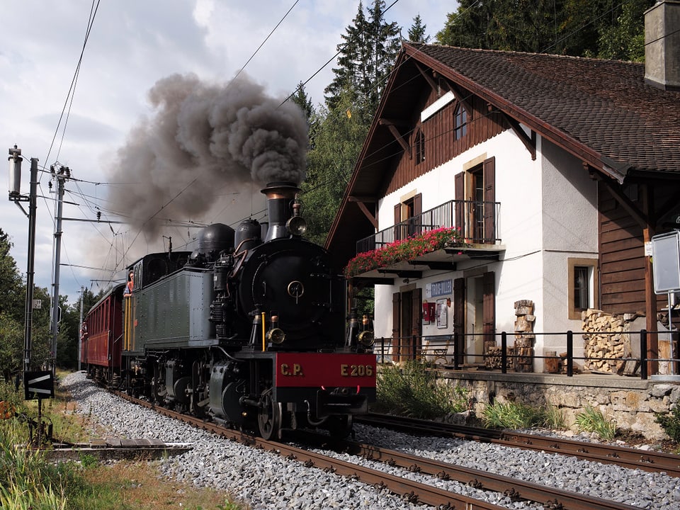 Eine alte, schwarze Dampflokomotive fährt rauchend an einem Haus vorbei.