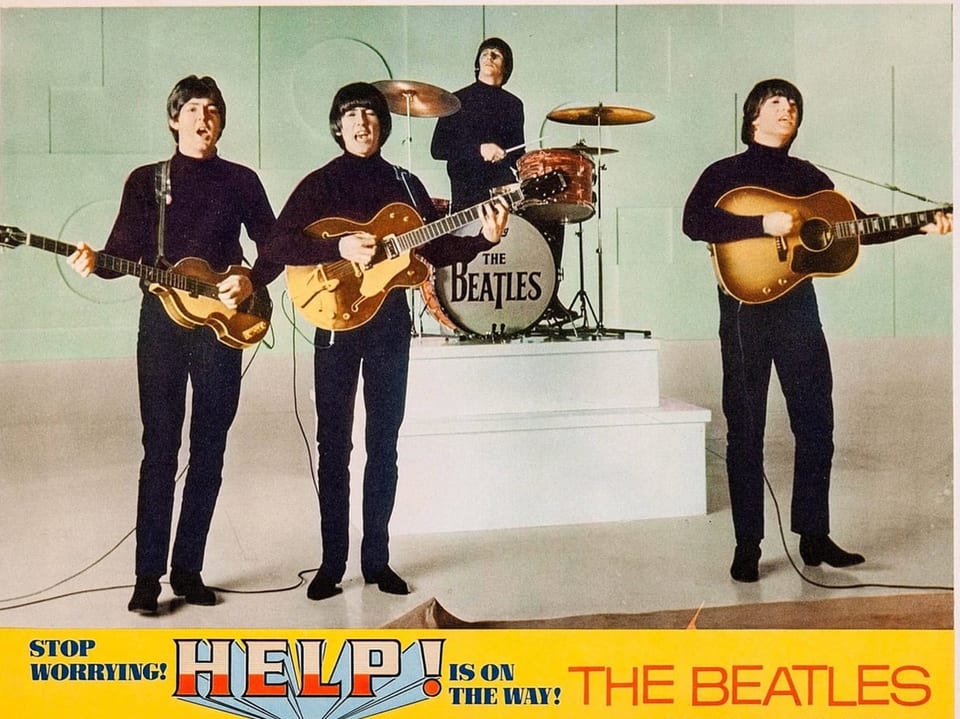 Die Beatles in schwarzen Rollkragenpullovern spielen Instrumente auf der Bühne mit dem Schriftzug 'HELP'.