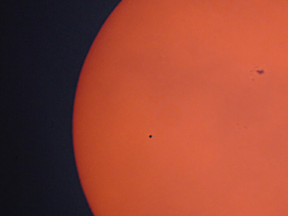 Ein winziger schwarzer Punkt wandert über die orange Fläche der Sonne. Rechts des Merkurs erkennt man noch einen Sonnenfleck.