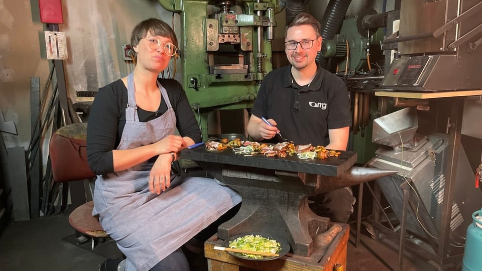 Frau mit Schürze und Mann mit schwarzem T-Shirt. Vor ihnen Essen auf einer Platte. 