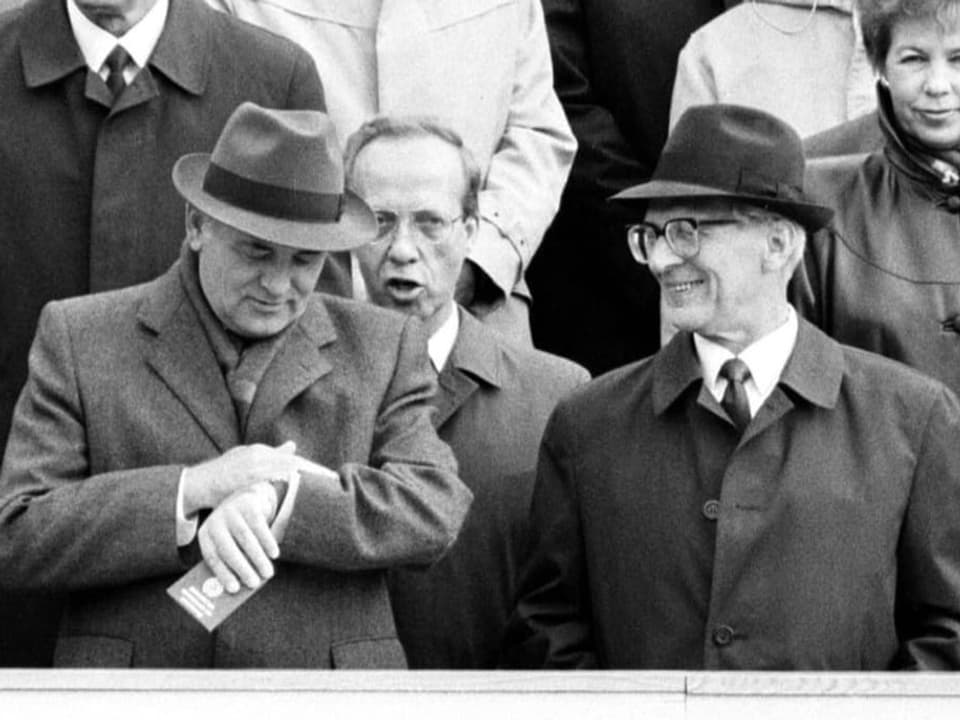 Zwei ältere Männer mit Mantel und Hut stehen auf einer Ehrentribüne.