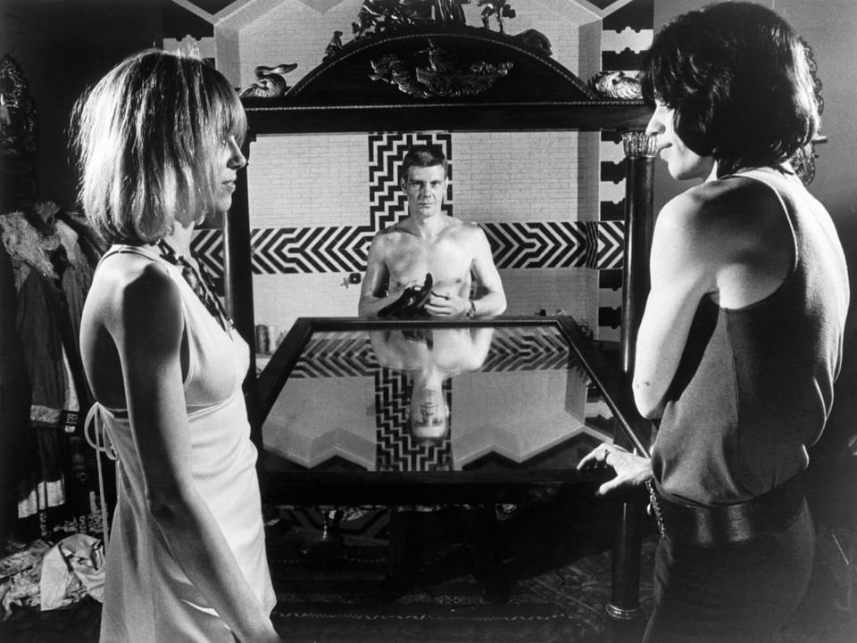 James Fox sitzt am Ende eines Glastisches und Anita Pallenberg und Mick Jagger schauen sich an.