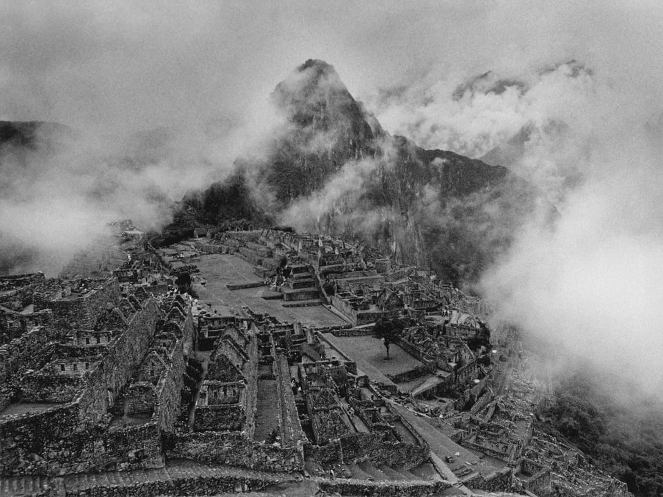 Schwarz-Weiss-Foto einer terassenartig angelegten Ruinenlandschaft vor einem Berg. Die ganze Landschaft ist von Wolken umgeben. 