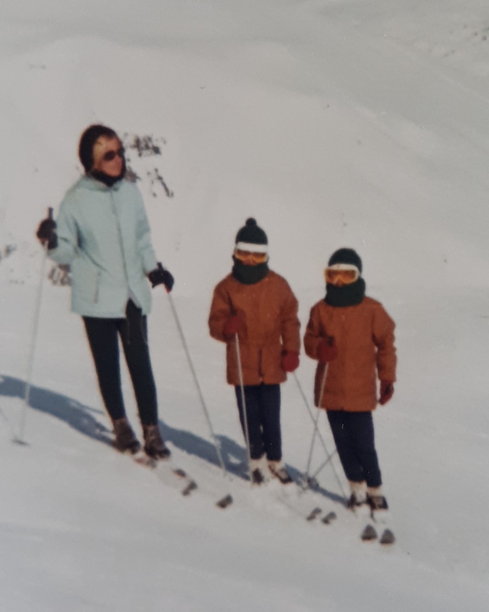 Eine Frau und zwei kleine Kinder auf Skis. Die Kinder tragen beide rote Jacken. 