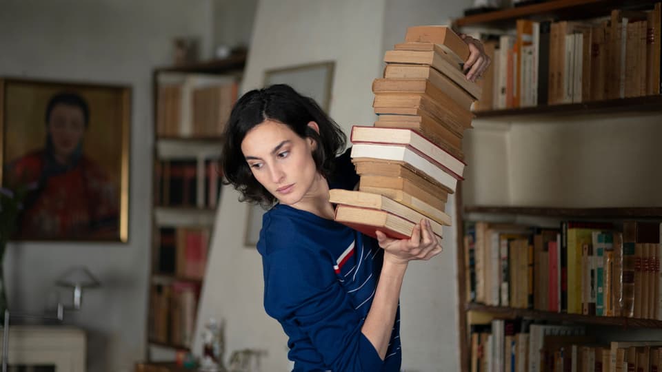 Eine Frau mit einem schiefen Bücherturm in der Hand.