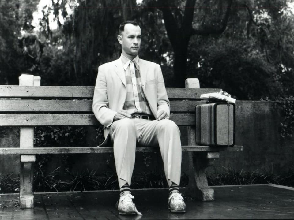 ZU sehen ist der Schauspieler Tom Hanks als Forrest Gump. Er sitzt auf einer Holzbank, seine Beine sind exakt parallel ausgerichtet. Er wartet augenscheinlich auf etwas.