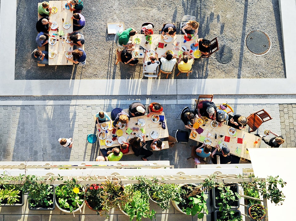 Blick von oben auf Pflanzen und Menschen, die an bunt bedeckten Tischen sitzen.