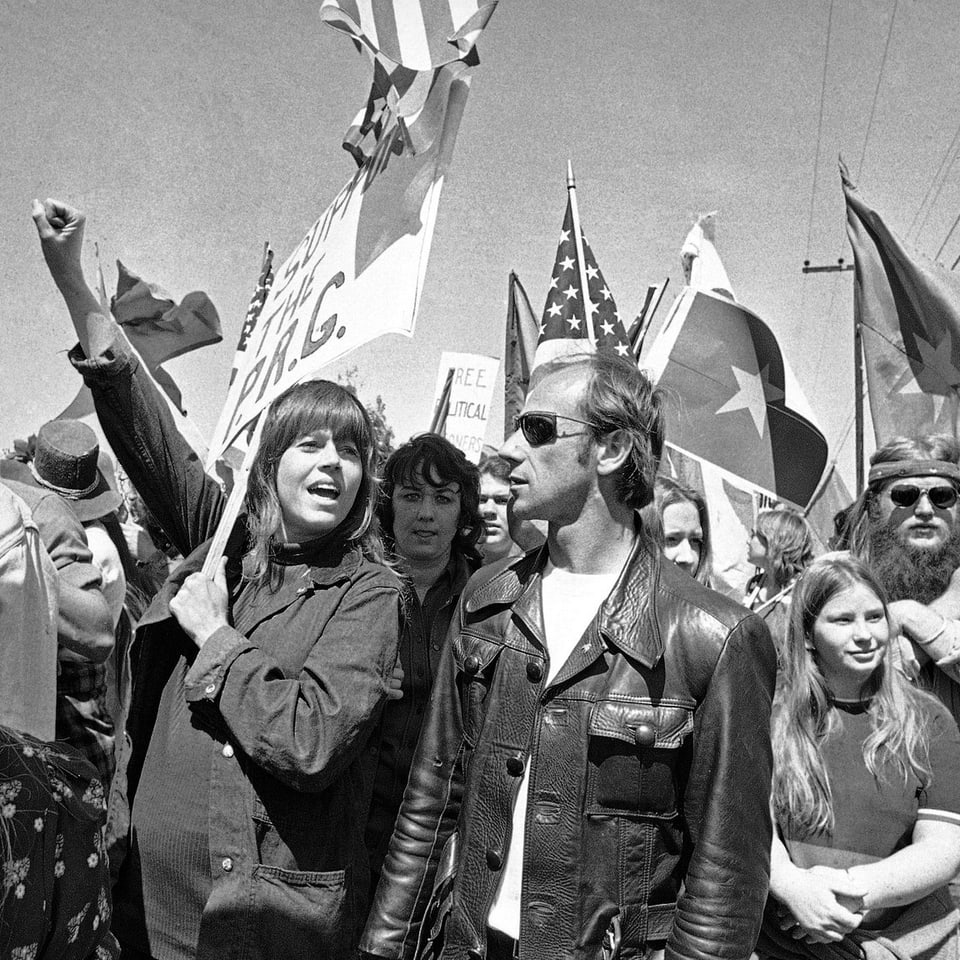Jane Fonda streckt die Faust in die Höhe, während hinter ihr sich ein Fahnenmeer offenbart. Hippies stehen um sie herum.