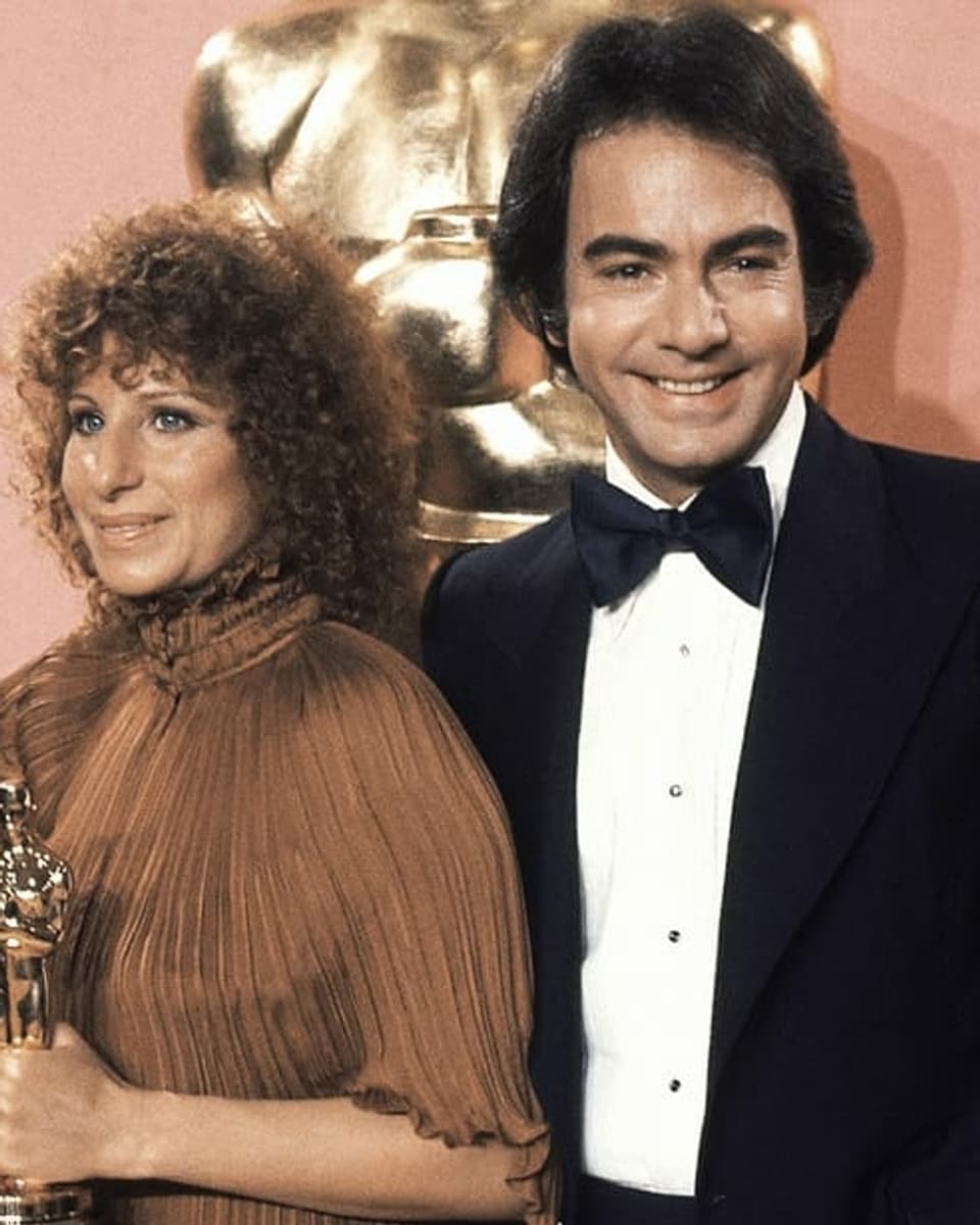 Barbra Streisand und Neil Diamonds posieren als Erwachsene lächelnd für die Kameras. Streisand hält ein goldenes Oscar-Männchen in der Hand. 