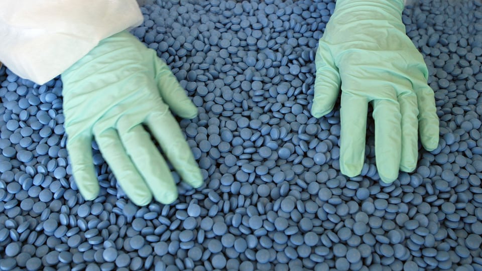Hände in Handschuhen durchmischen einen Berg von blauen Tabletten.