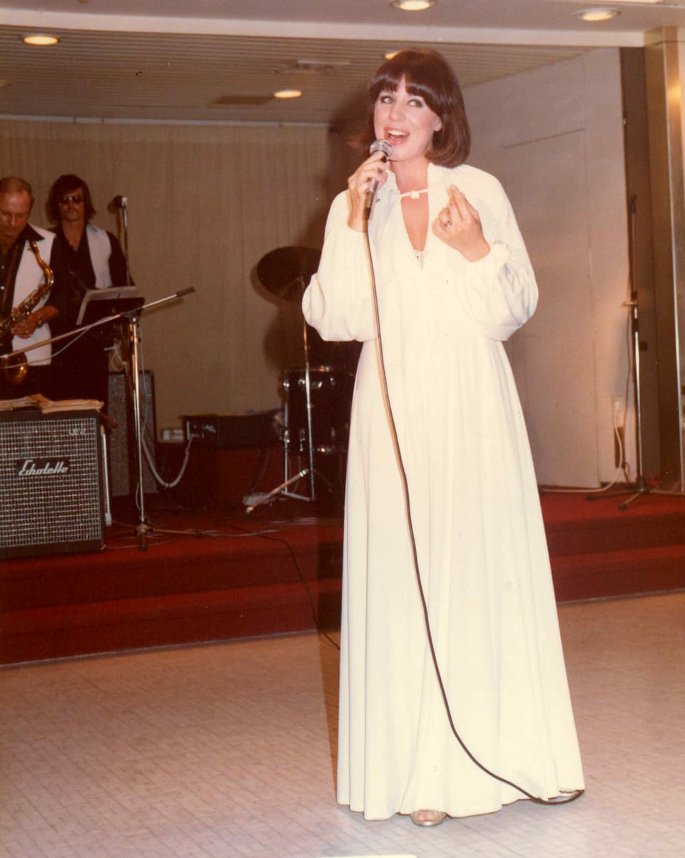Sängerin während Auftritt mit Band im Hintergrund. Sie trägt ein langes, weisses Kleid. 