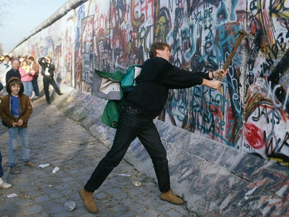Jüngerer mann in schwarzer Kleidung schwingt einen Hammer und haut auf eine von Graffiti überzogene Mauer ein.