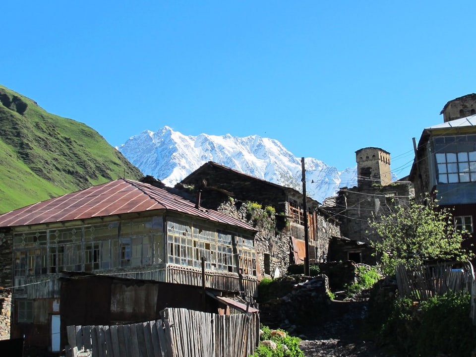Ein Foto von Ushguli: Im Vordergrund sieht man ein paar Häuser, hinten einen Berg und blauen Himmel. 
