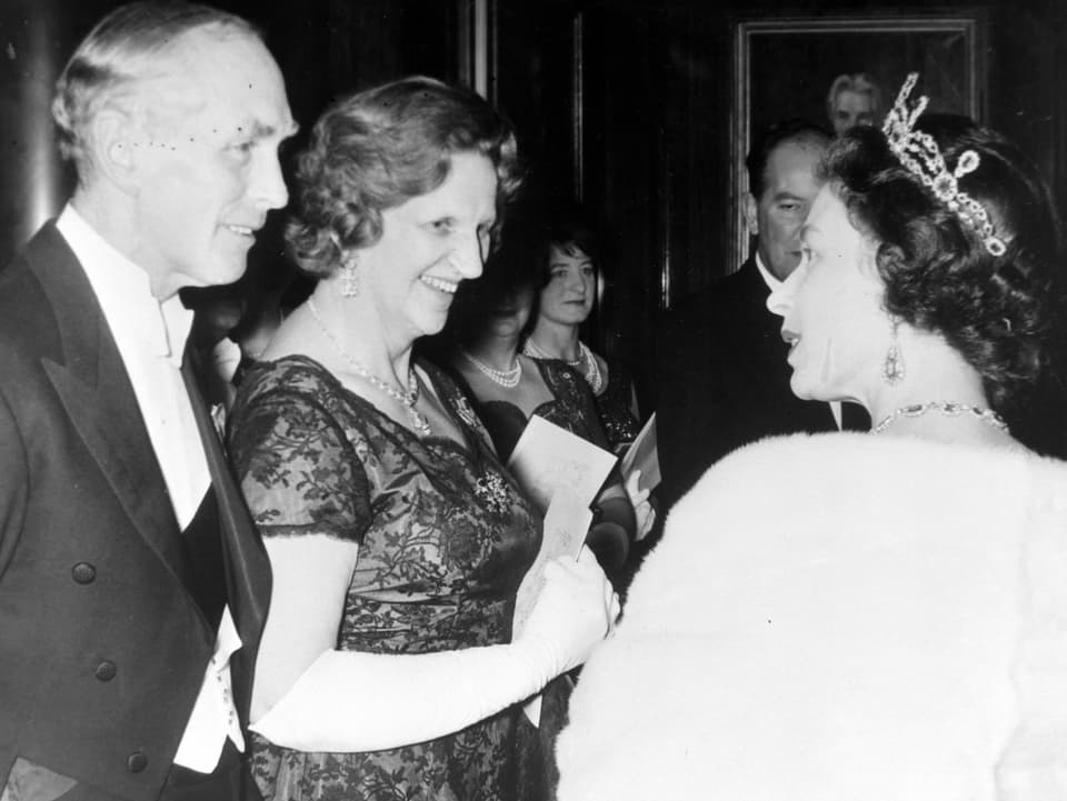 Aufnahme in schwarz-weiss. Menschen in festlicher Kleidung. Ein Ehepaar begrüsst die Queen, die eine Krone auf dem Kopf trägt.