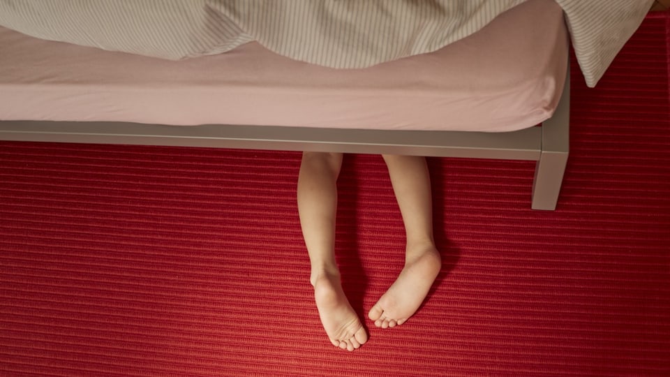 Kinderfüsse ragen unter einem Bett hervor. Das Kind versteckt sich vor möglicher Gewalt durch Bezugspersonen.