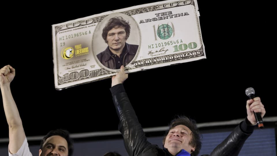 Milei hält ein Pappbild eines 100-US-Dollar-Scheins mit seinem Gesicht in die Höhe.