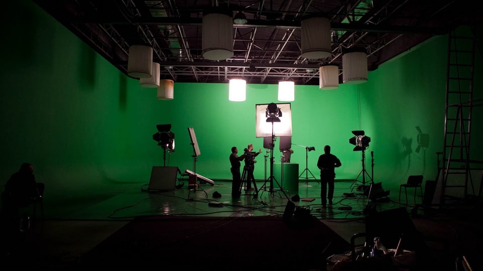 Ein Studio mit Lampen und Greenscreen. In der Mitte stehen drei Menschen.