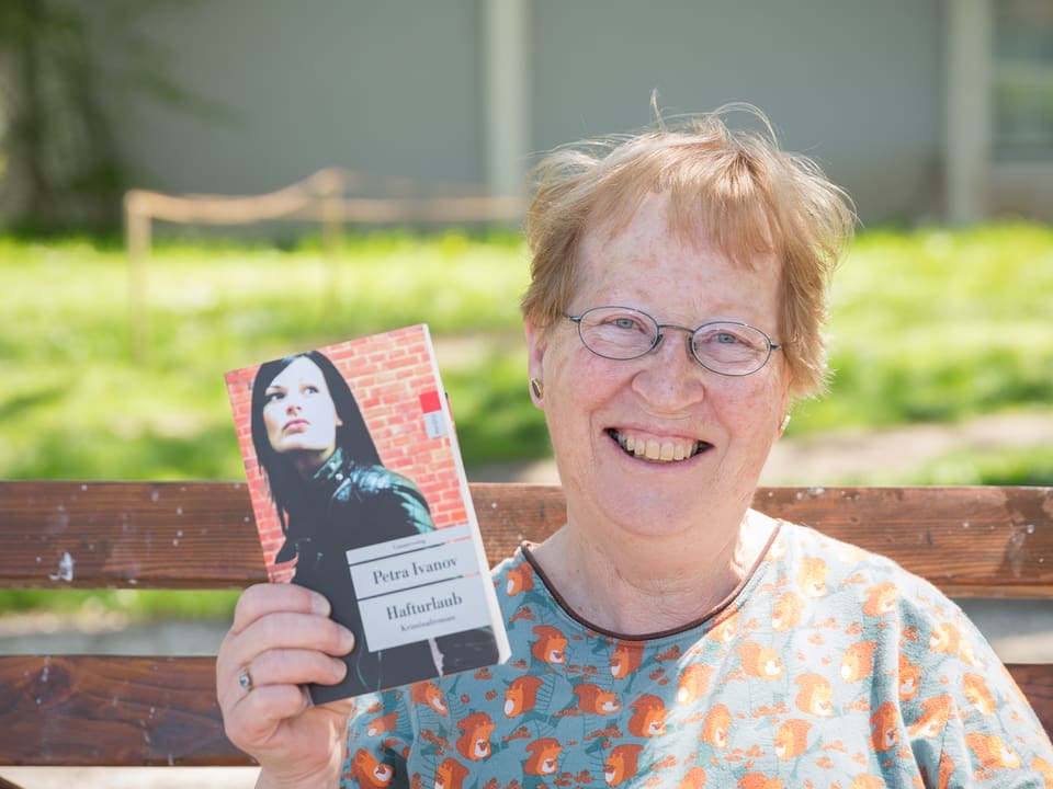 Eine Frau mit Brille sitz lachend auf einer Parkbank und hält ein Buch in die Höhe.
