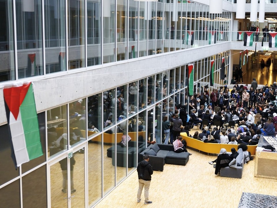 Menschen in einer modernen Lobby mit Glasfassade und hängenden Flaggen.