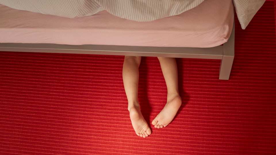 Symbolbil: ein Kind unter einem Bett, man sieht nur die nackten Beine.