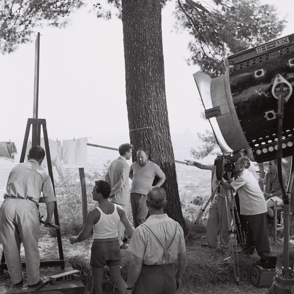 Eine Szene unter einem grossen Baum. Im Vordergrund rechts steht eine grosser Filmscheinwerfer. In der Mitte des Bildes sind zwei Schauspieler vor einem Baum, der eine ist daran angelehnt. Neben ihnen steht die Kamera.