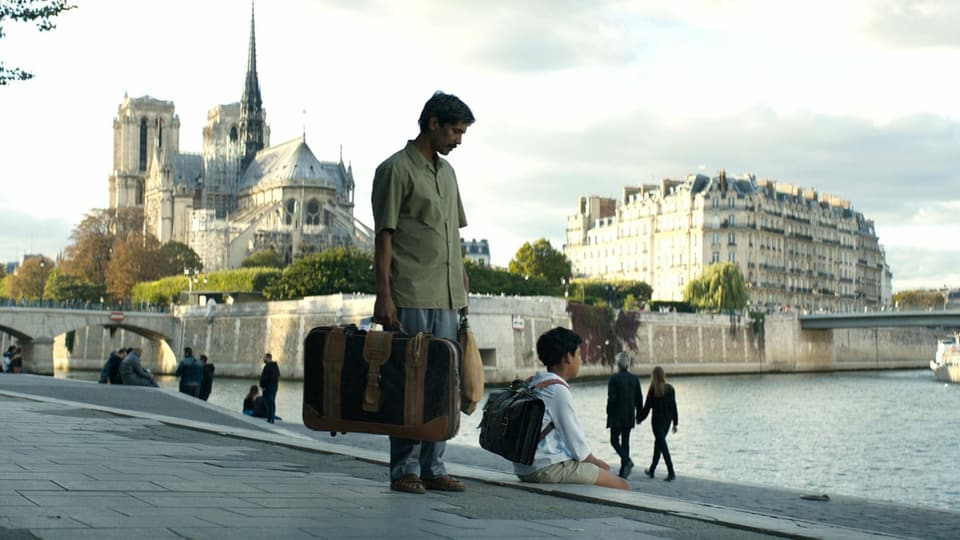 Ein Vater mit sienem Sohn befinden sich mit Koffer an der Seine in Paris. 