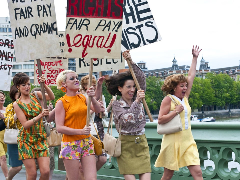 Frauen im schrillen 60er-Jahre-Look demonstrieren mit Plakaten auf einer Brücke.