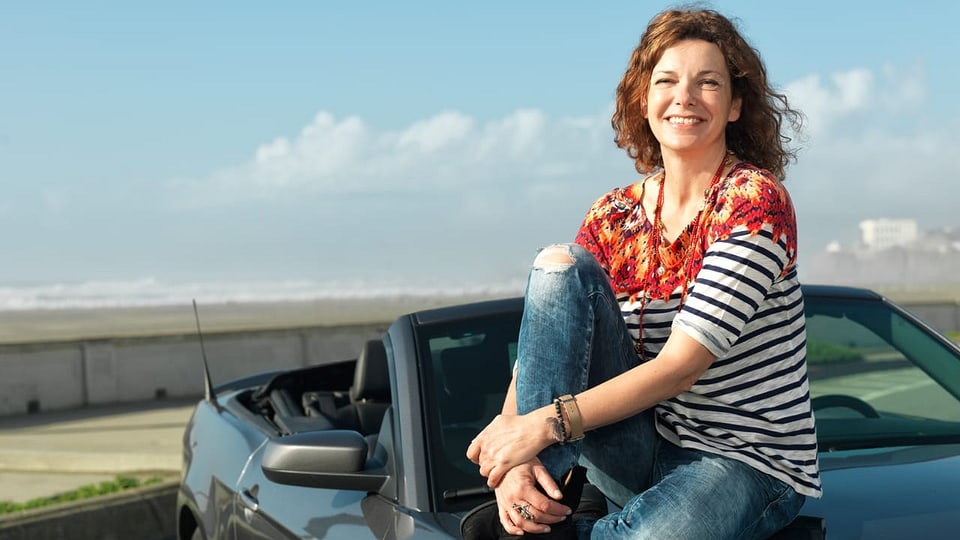 Eine Frau mittleren Alters sitzt auf einer Kühlerhaube eines Autos.