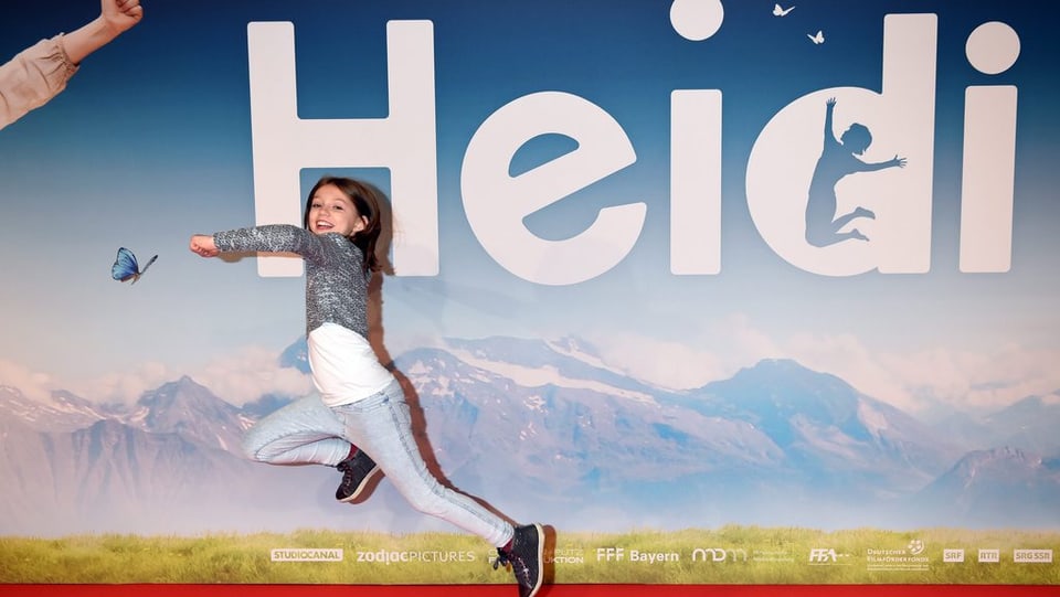 Heidi-Hauptdarstellerin Anouk Steffen springt vor einem Heidi-Plakat in die Höhe.