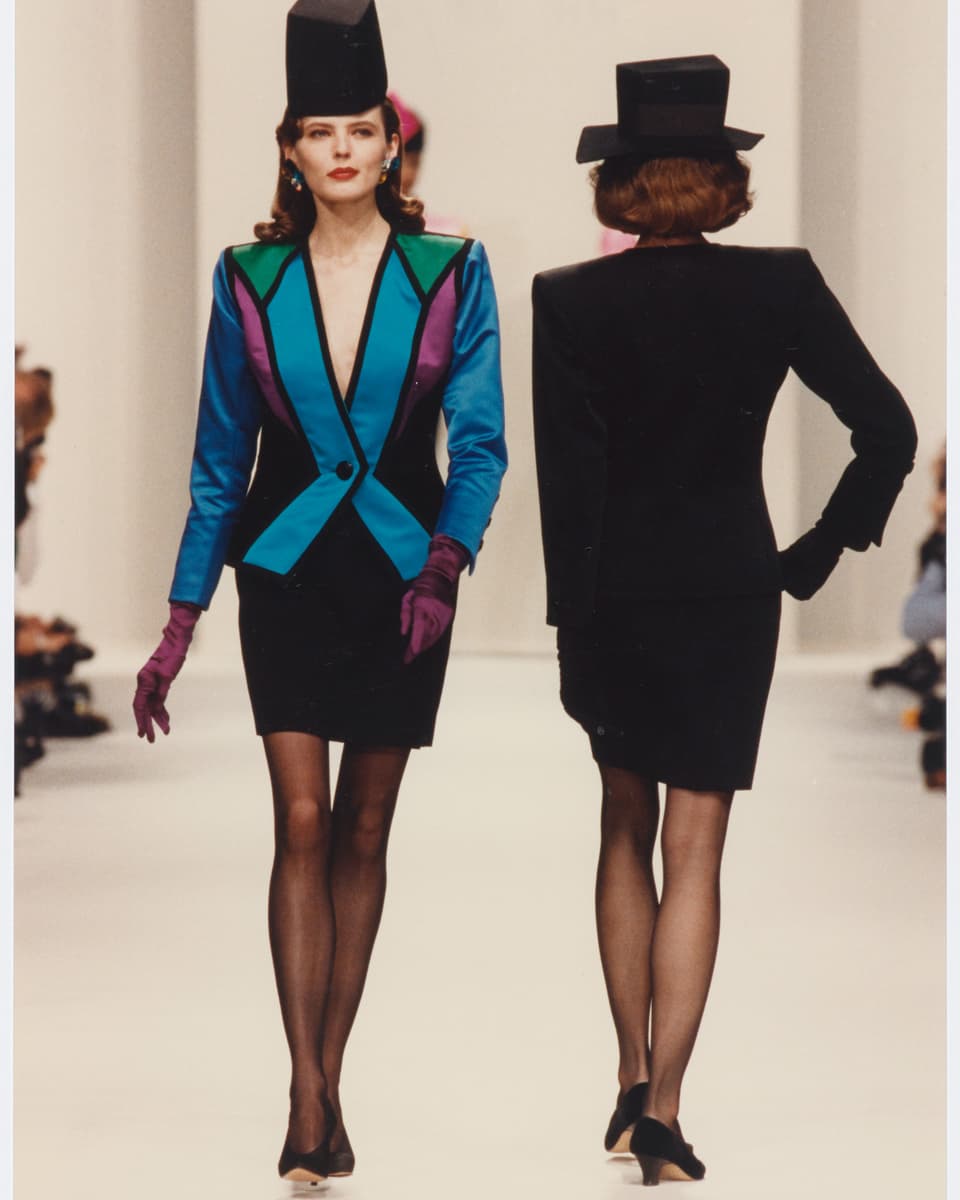 two women on catwalk, one facing front, wearing blazer of blue, black and purple and black skirt.