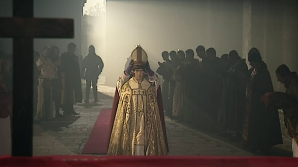 Frau im weissgoldnen Paps-Umhang, wird gekrönt, steht auf rotem Teppich, in einer Reihe rechts und links Männer.