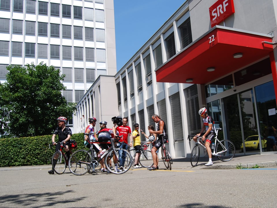 Zu sehen ist eine Gruppe Velofahrer, die neben ihren Velos stehen. Vor dem Radiostudio.