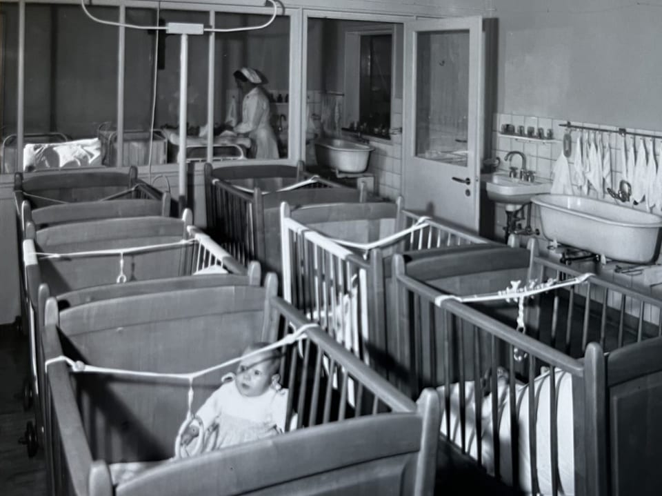 Historisches Bild von einem Krankenhaus-Säuglingszimmer mit mehreren Babybetten und einer Krankenschwester.