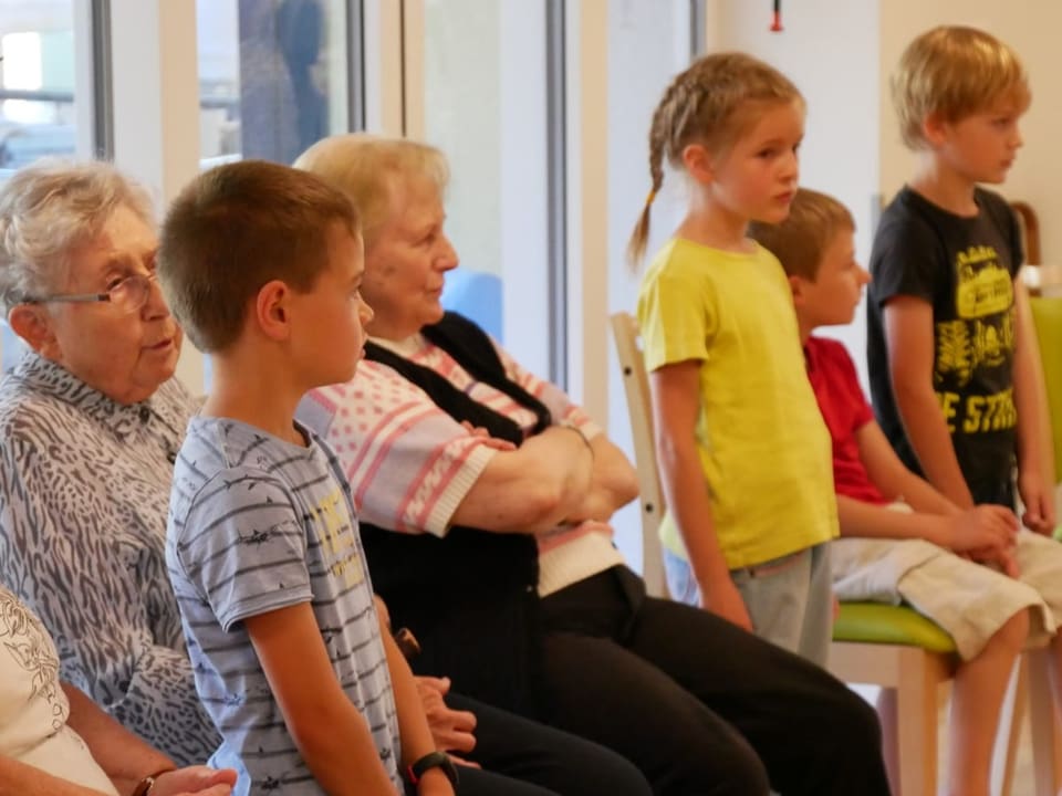 Kinder und zwei Seniorinnen sitzen bzw. stehen nebeneinander