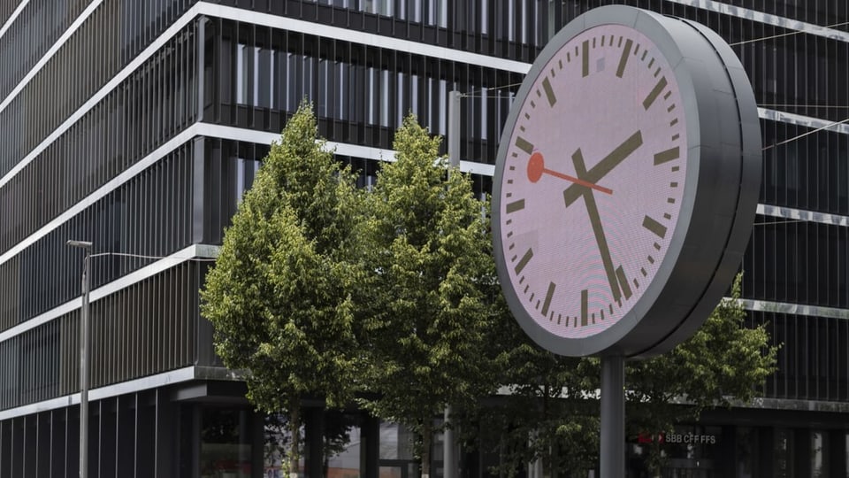 Sehr grosse Uhr steht vor einem Gebäude.