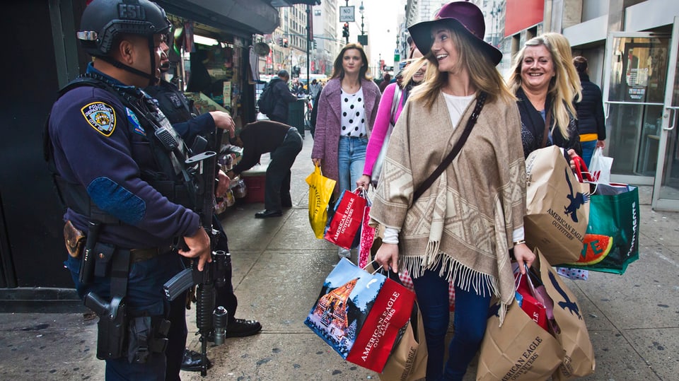 Zwei Frauen bepackt mit Einkaufstaschen gehen auf die Kamera zu und blicken auf zwei Polizisten links im Bild