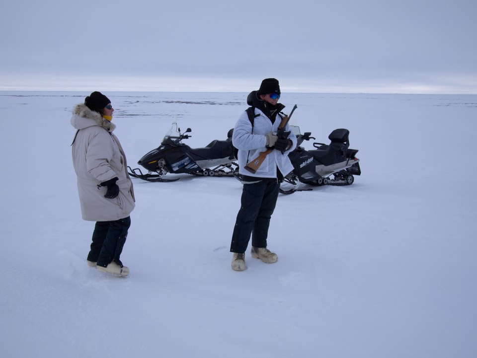 Zwei Personen mit einem Gewehr auf einer Eisfläche.