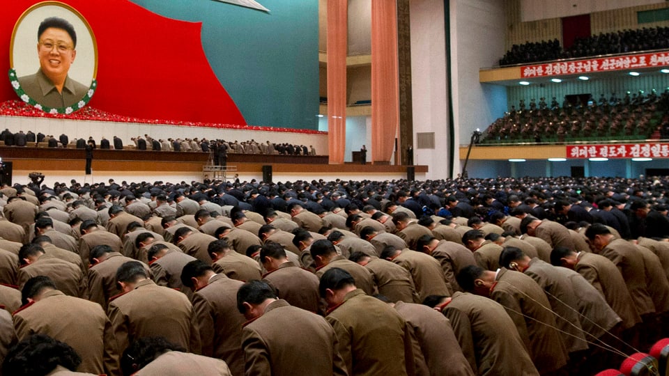 Staatsangestellte und Armeeangehörige verneigen sich in einem grossen Saal, an der Wand hängt ein Portrait von Kim Jong Il.