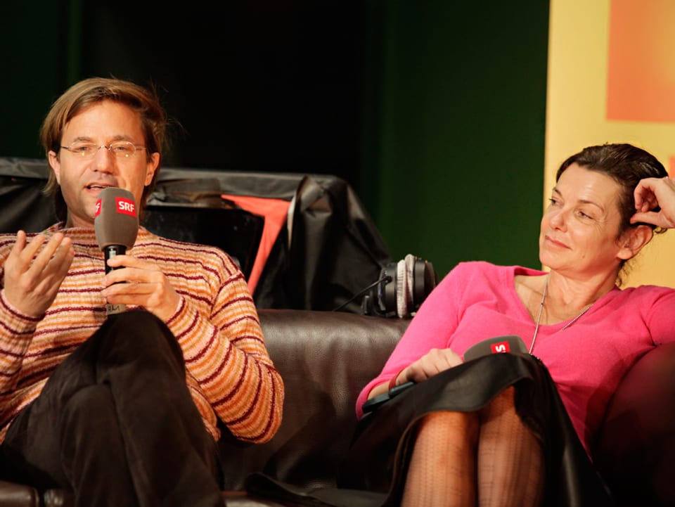 Ein Mann mit Mikrofon sitzt neben einer Frau auf einem Sofa.