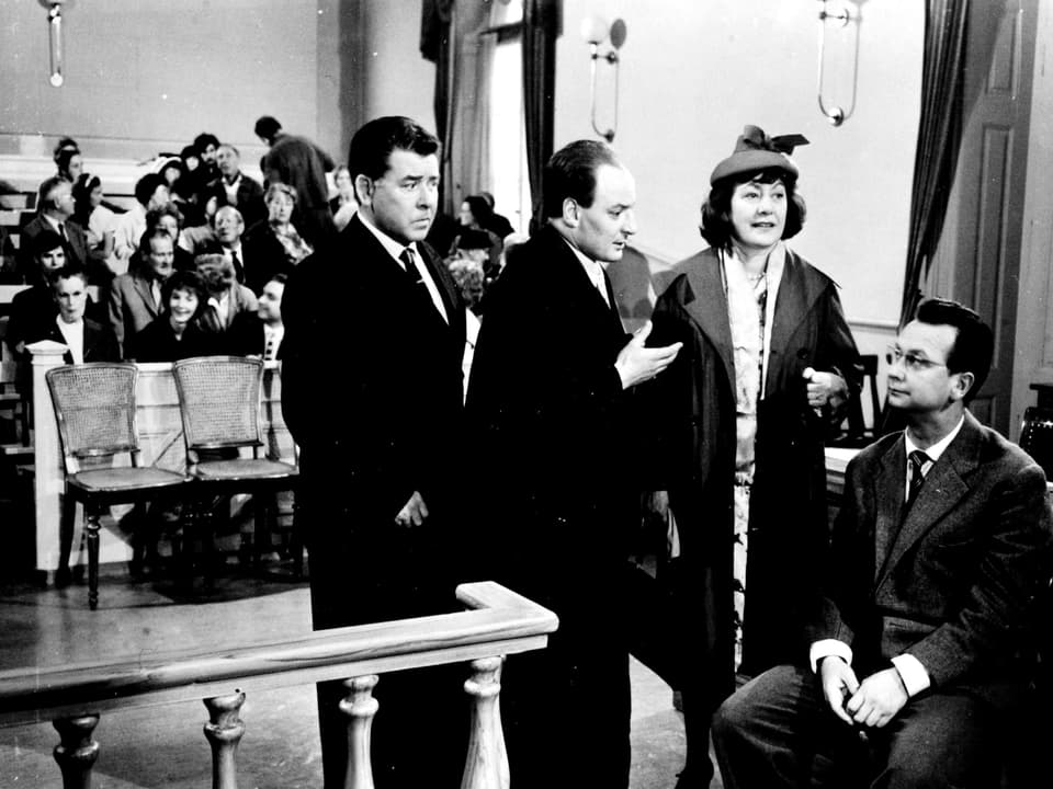 Drei Männer und eine Frau diskutieren in einem Gerichtssaal. Im Hintergrund sitzen Menschen und verfolgen die Diskussion.