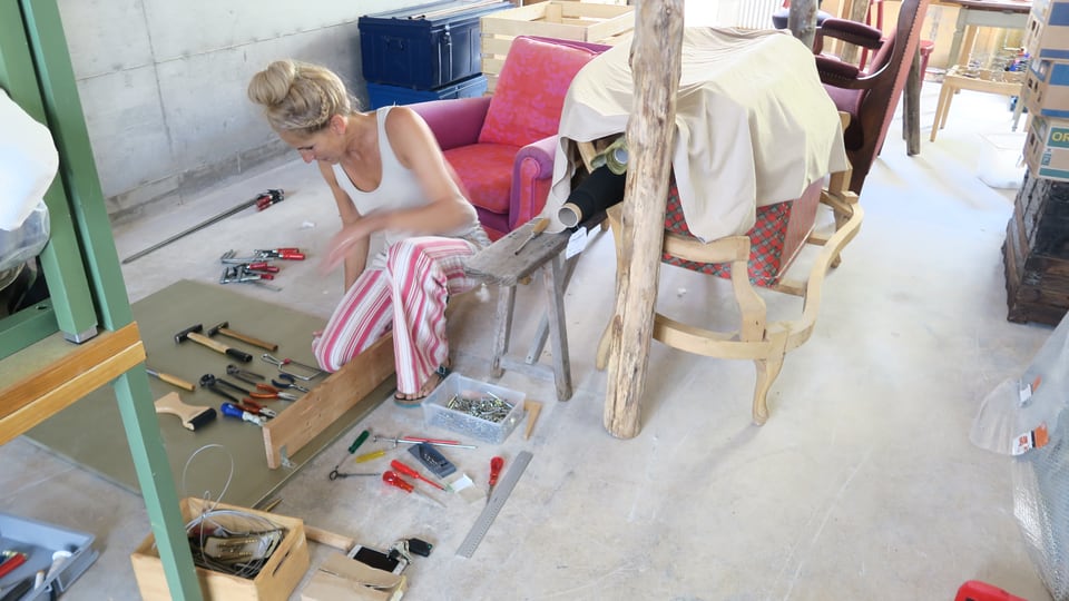 Eine Frau in einem Atelier kauert am Boden, vor sich diverse Werkzeuge. 