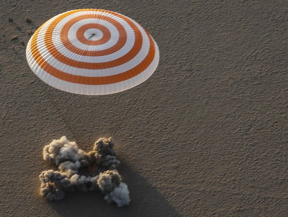 Raumkapsel landet im Sand, rot-weisser Fallschirm.