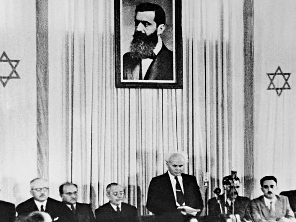 An einem langen Tisch sitzen Männer, dazwischen steht David Ben-Gurion, der erste Premierminister. Über ihm ein Bild von Theodor Herzl.